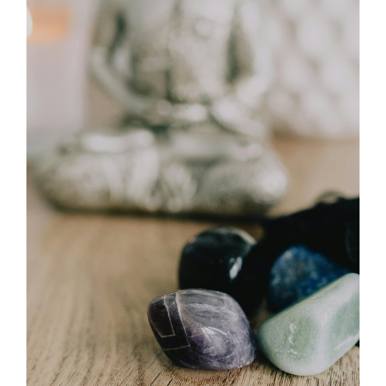 A close-up of polished tumbled stones including amethyst, aventurine, and sodalite, resting on a wooden surface with a blurred silver Buddha statue in the background, evoking a peaceful, meditative energy.