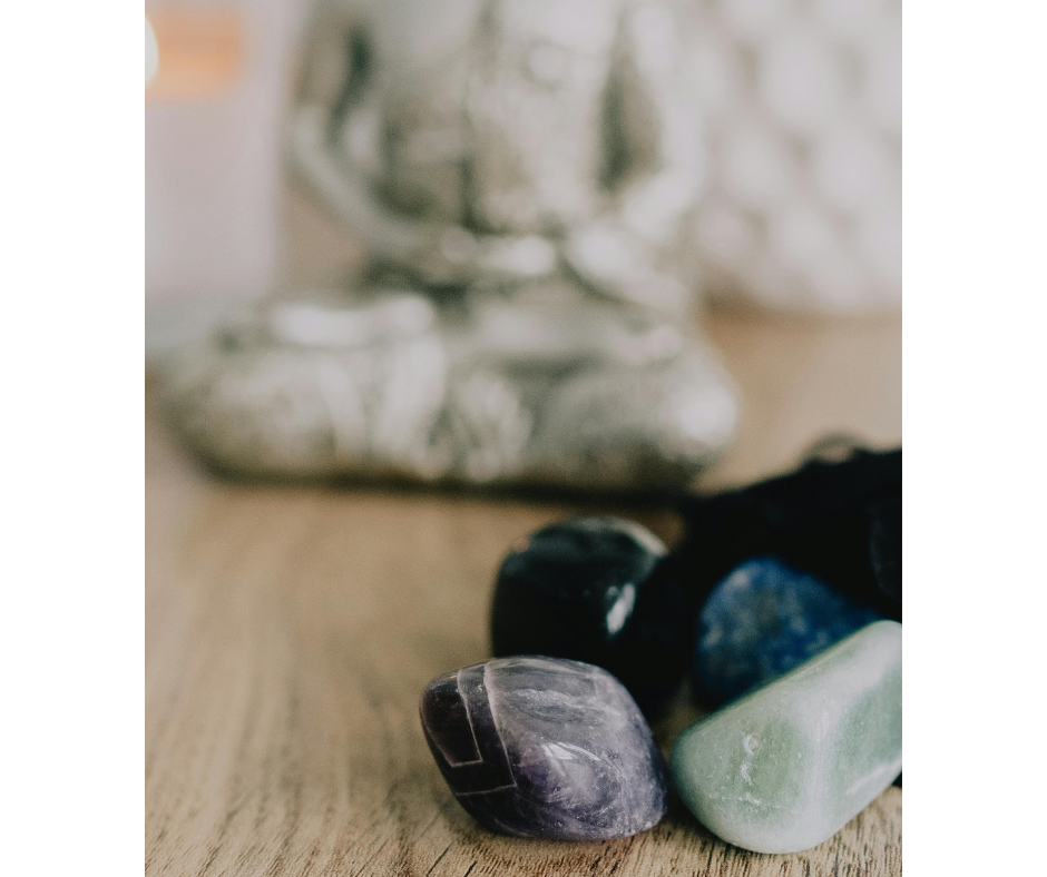 A close-up of polished tumbled stones including amethyst, aventurine, and sodalite, resting on a wooden surface with a blurred silver Buddha statue in the background, evoking a peaceful, meditative energy.